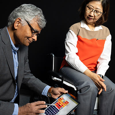 An adult seated in a wheelchair looks toward a tablet held by another adult, displaying a colorful grid of pressure‑mapping data against a dark background.
