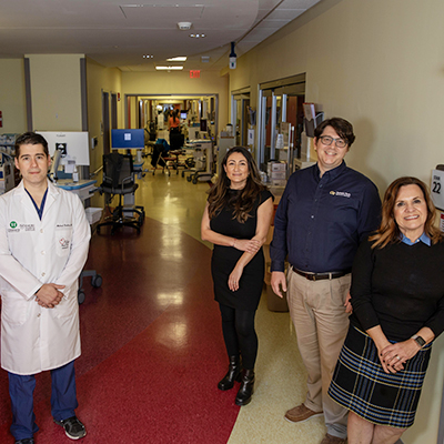 Four adults stand in a hospital hallway lined with medical equipment and workstations, with additional staff and activity visible farther down the corridor.

