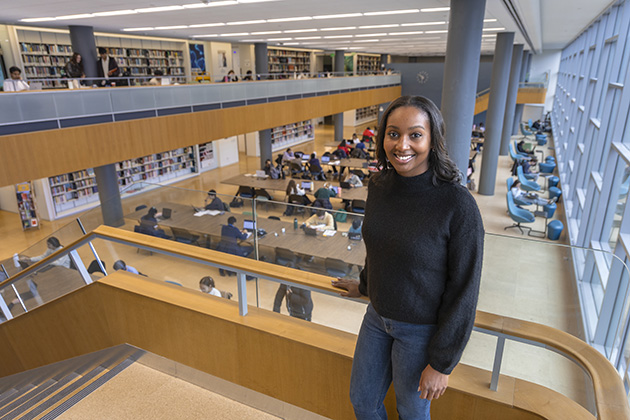 Person standing on an indoor staircase overlooking a spacious library with rows of bookshelves and people studying at tables.