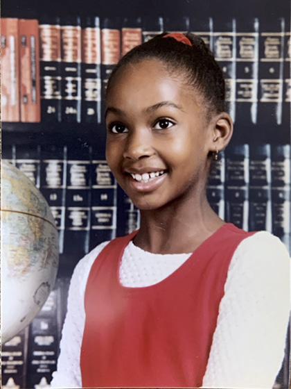 Child wearing a red jumper over a white shirt, posing next to a globe with a backdrop of encyclopedias.