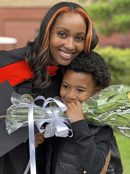 Graduate in a black gown with a red stole holding a bouquet of flowers, standing close to a child dressed in black.