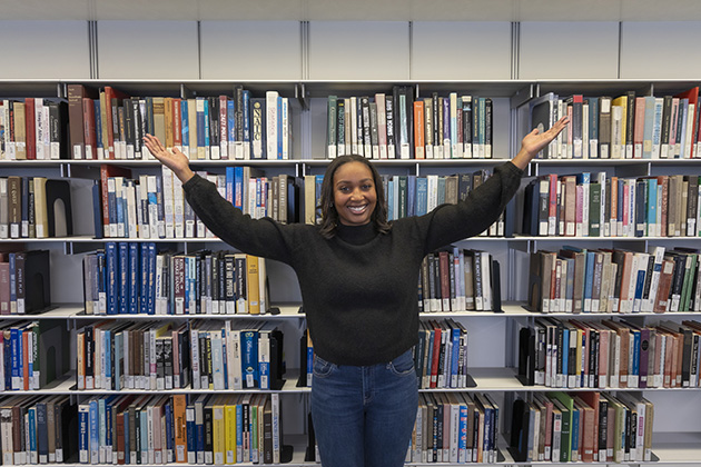 Person standing in front of a large bookshelf filled with books, arms raised outward in a welcoming gesture.