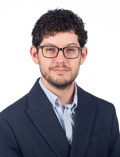 Person dressed in a dark blazer and light blue collared shirt posing against a plain white background