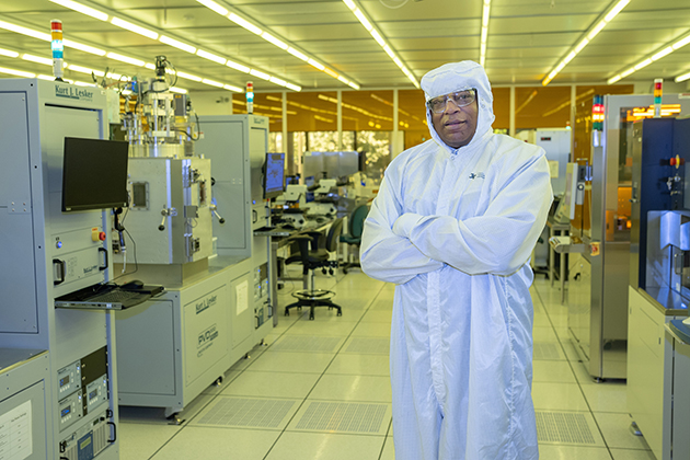 A person in a full cleanroom suit stands with arms crossed in a brightly lit semiconductor fabrication lab with equipment in the background.