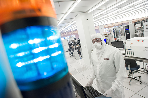 A cleanroom technician in protective gear works at a computer workstation in a semiconductor lab, with a blue signal light in the foreground and lab equipment behind them.
