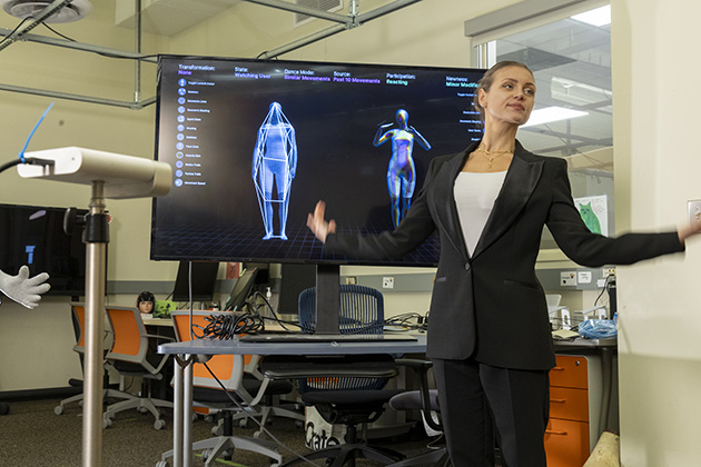 female with her hair pulled up and wearing a black suit jacket standing in front of two screen monitors showing blue outlines of human forms
