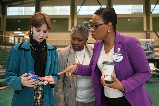 Three people standing together at an event, examining a small model race car while one person gestures and holds a beverage cup.