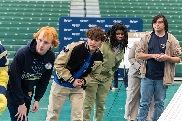 Students leaning forward and observing an activity together in an indoor gymnasium during a Georgia Tech event.