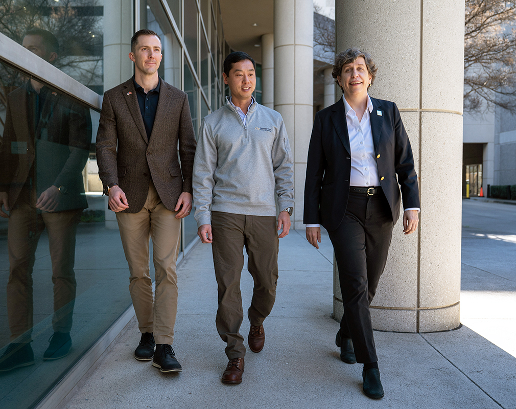 Three people walking together outside a modern building with large glass windows and concrete columns.