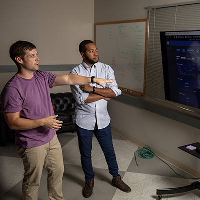 Two people standing in a workshop area reviewing notes and tasks on a large whiteboard with a grid layout.
