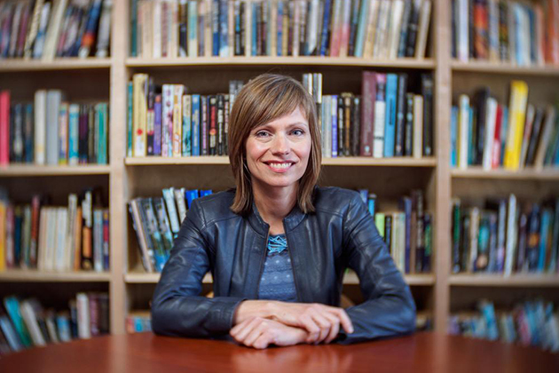 Person seated at a wooden table wearing a black leather jacket with bookshelves filled with books in the background