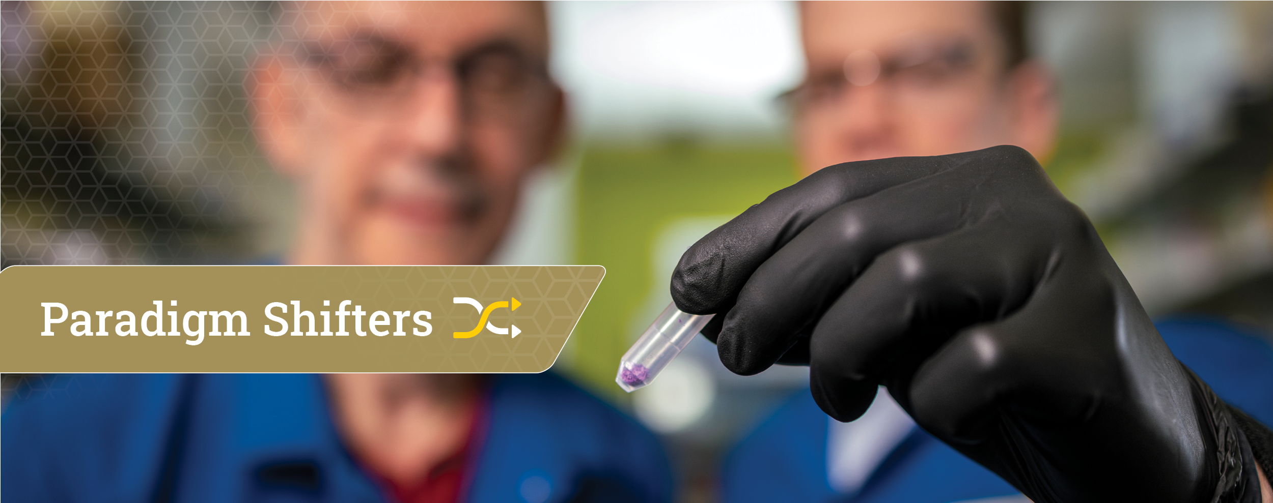 A gloved hand holds a small vial with purple liquid in front of two people wearing blue lab coats, with a “Paradigm Shifters” banner partially visible on the left side of the image.