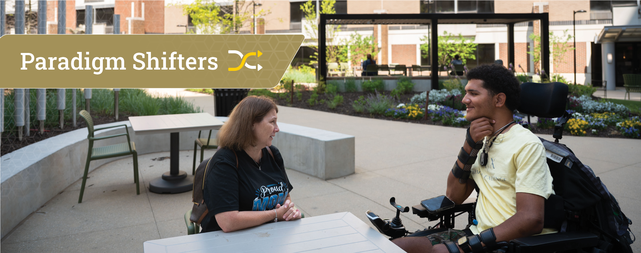 Two people seated at an outdoor campus table in conversation, one using a powered wheelchair with assistive controls, in a landscaped courtyard setting.
