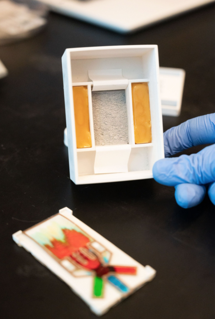 The new user-friendly, disposable liquid flow assay includes a heating unit (being held) and the paper-based assay ("dipstick") with built-in flow controller (loaded with dye solutions to for demonstration purposes), showing the sequential release of different fluids to the reaction spots.   Photo by Allison Carter