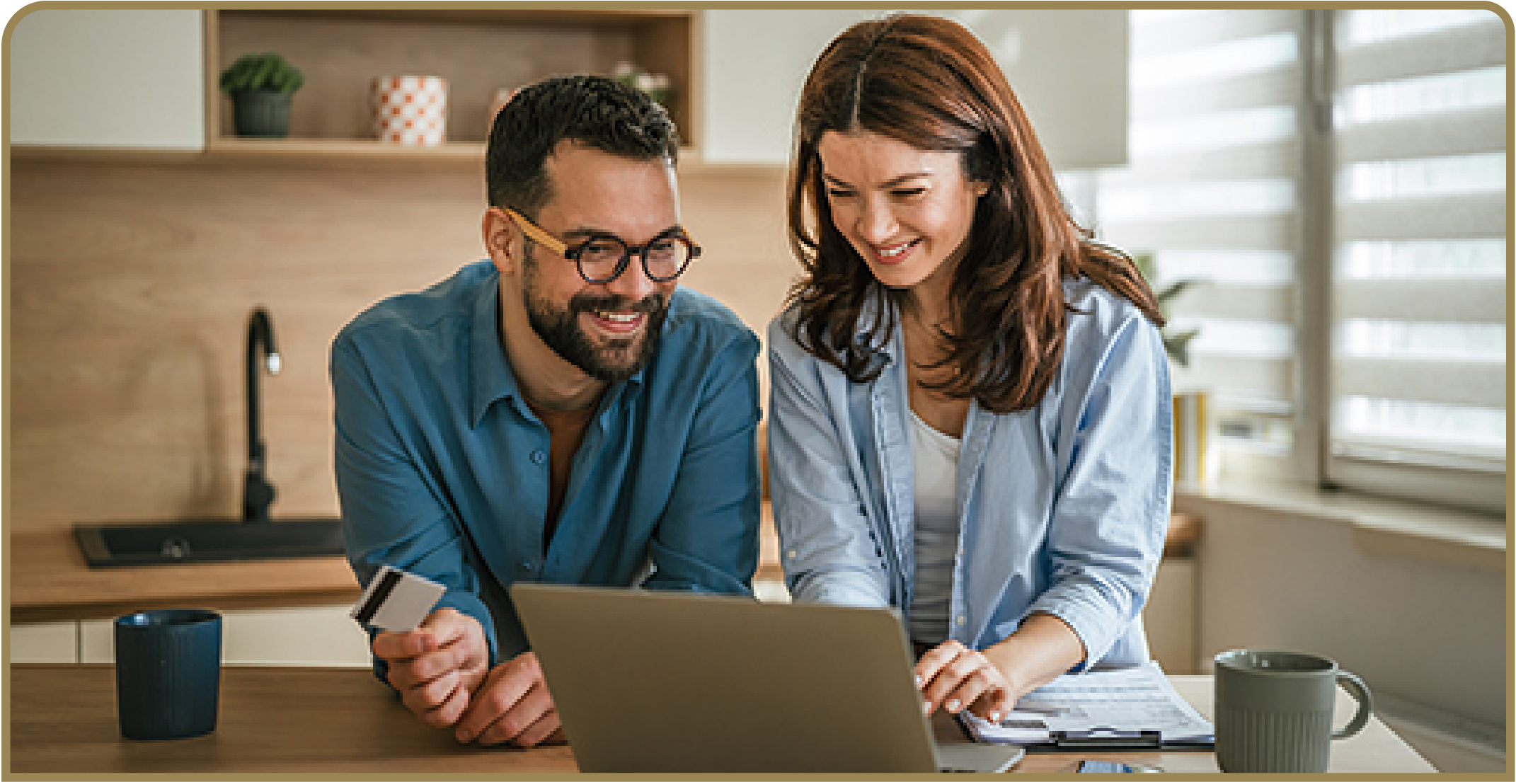Two people review information on a laptop while one holds a credit card and the other references printed documents at a table..