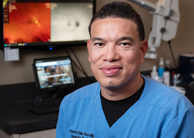 Person in blue medical scrubs seated at a desk with computer monitors showing clinical images and equipment in the background.