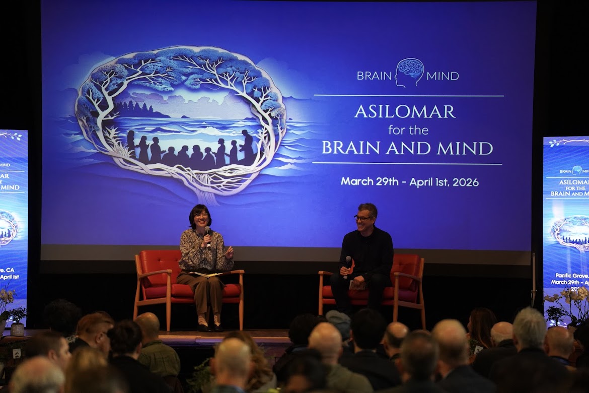 Seated on the left, Karen Rommelfanger speaks on a panel at the 2026 Asilomar for the Brain and Mind conference. Panelists sit on stage in front of a large screen displaying the conference name, dates, and a brain-themed graphic, with an audience visible in the foreground.