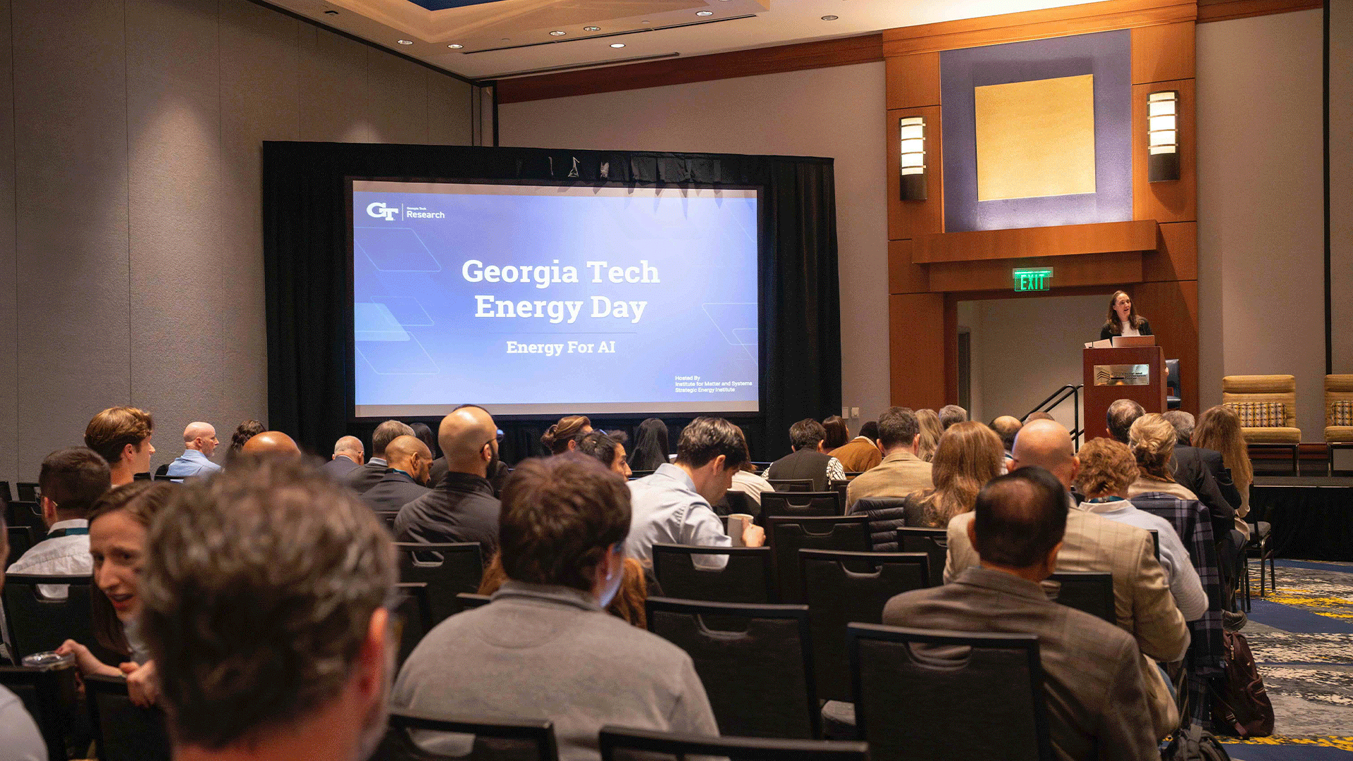 A wide view of a conference room shows attendees seated and facing a stage with a large screen reading “Georgia Tech Energy Day: Energy for AI.” Marta Hatzell stands at a podium to the right of the screen.