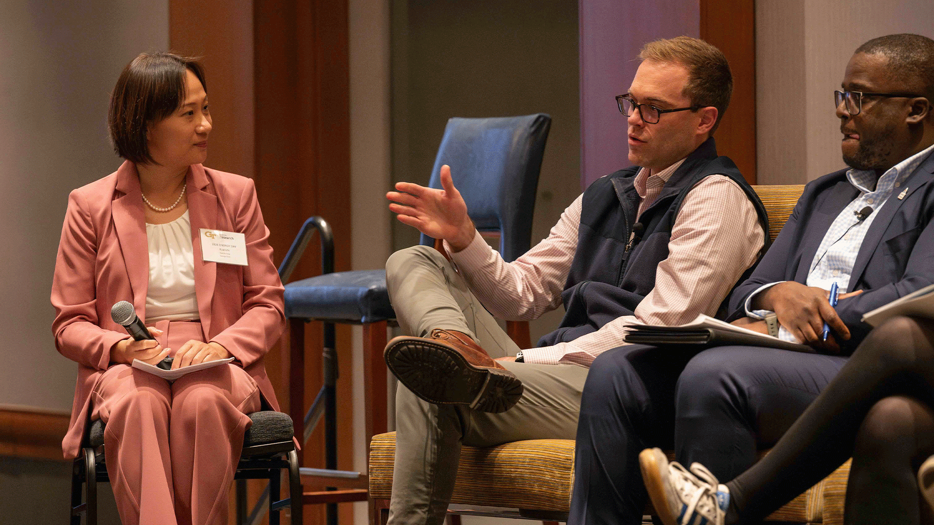 Three panelists sit on stage during a discussion, with one man gesturing as he speaks while the others listen. The moderator holds a microphone and looks toward him.