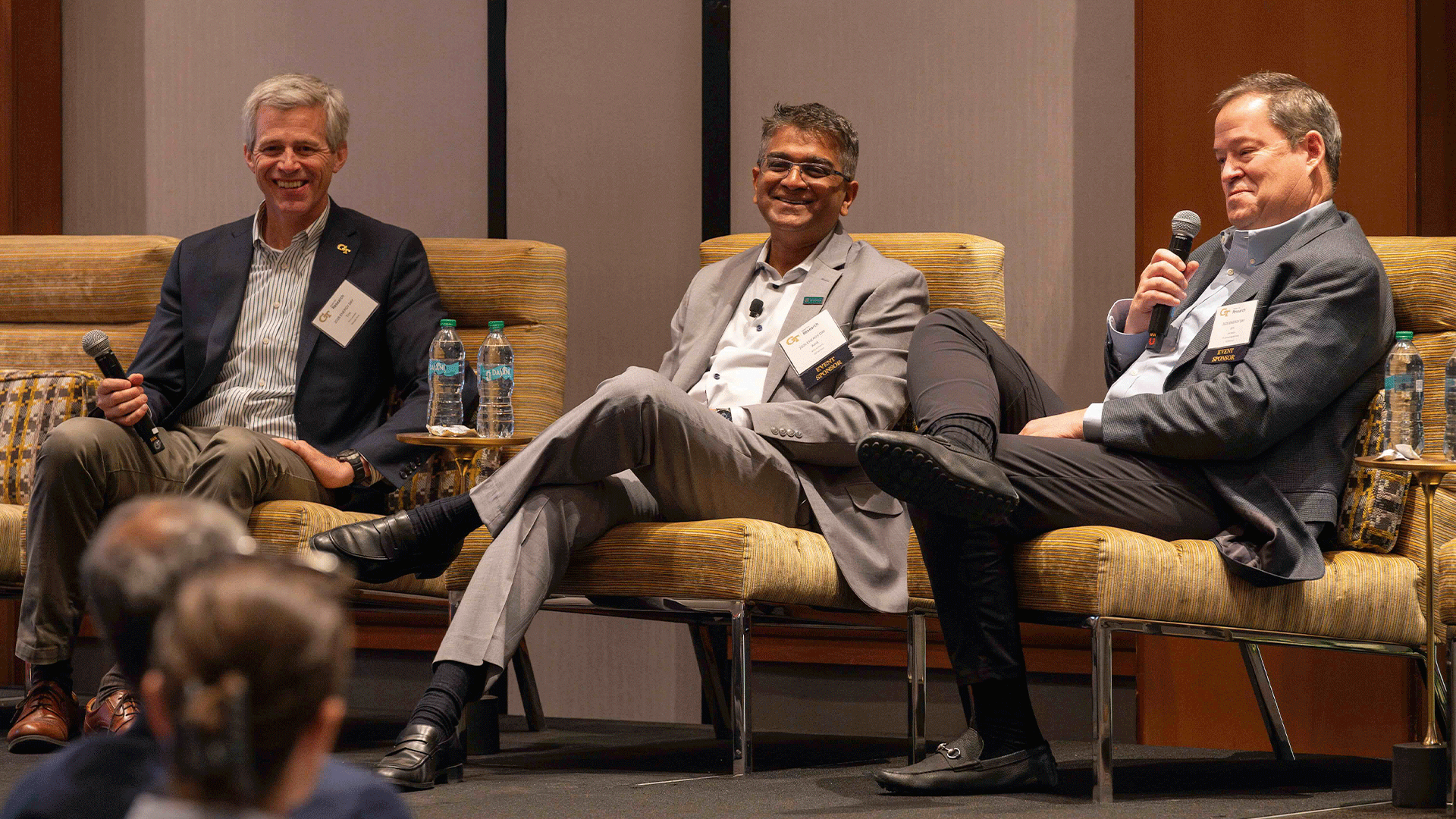 Three men sit on stage in a panel discussion, smiling and holding microphones. Water bottles rest on small tables beside their chairs.