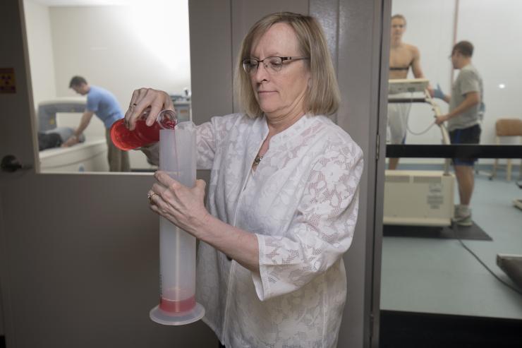 <p>Professor Mindy Millard-Stafford researches in exercise physiology, including studies on how dehydration and exertion can reduce physical and cognitive performance. In the background, a volunteer lies on a device that measures body composition (l.) and another volunteer sweats out fluids under exertion in a heated chamber (r.). Millard-Stafford is a past president of the American College of Sports Medicine. Credit: Georgia Tech / Christopher Moore</p>