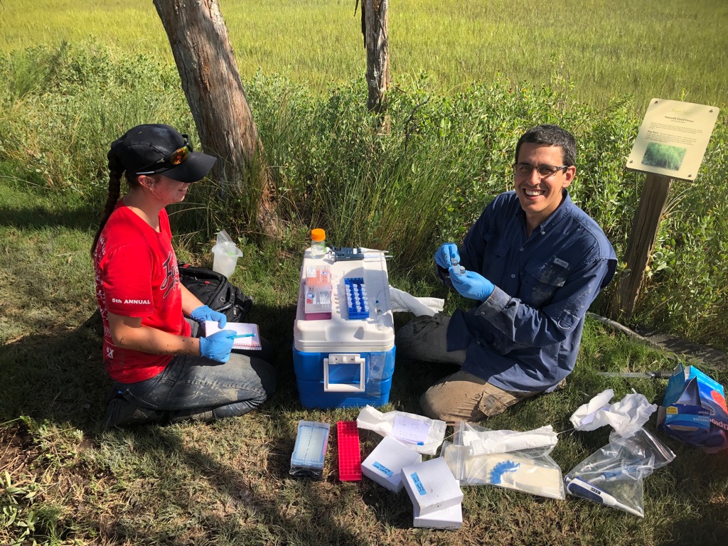 Two people sitting on a ground with a cooler and scientific equipment (including sample vials) between them. 