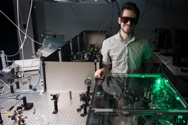 <p>Graduate research assistant Felix Thouin stands at a protective laser casing in the lab of Carlos Silva, in which laser light is adjusted and filtered to desirable qualities in the visible range. Behind Thouin, visible range laser light undergoes more processing to ready it for use in measuring materials qualities. Credit: Georgia Tech / Allison Carter</p>
