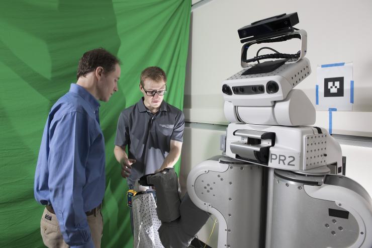 <p>A PR2 robot puts a gown on Henry Clever, a member of the research team.</p>