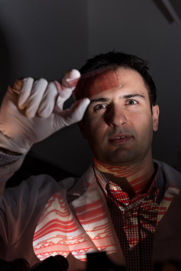 <p>Fatih Sarioglu, an assistant professor in the Georgia Tech School of Electrical and Computer Engineering, is shown examining a microfluidic device designed to capture cancer cell clusters. An image of the chip is projected onto his shirt. (Credit: Rob Felt)</p>