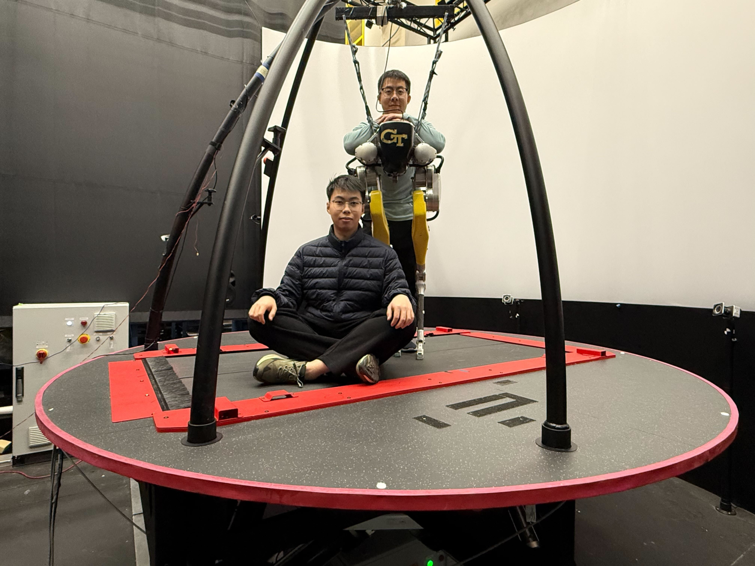Two individuals on a large circular motion platform in a research lab, with one person seated cross-legged on the platform and another suspended in a harness wearing a Georgia Tech exoskeleton system.