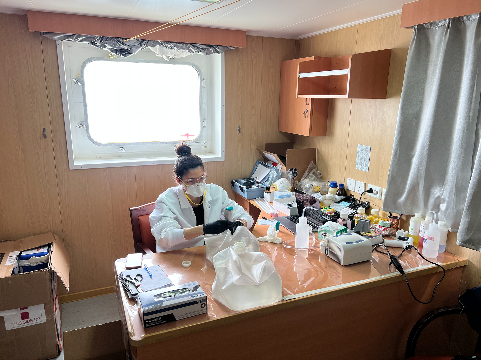 A woman in a lab coat sits at a desk in a ship cabin. She is surrounded by bottles and scientific measurema