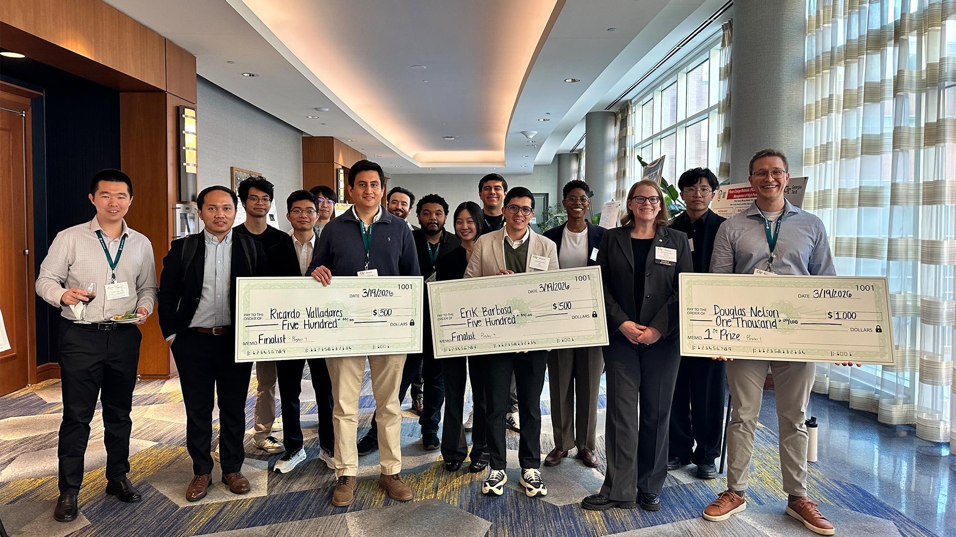 A group of people stand indoors at an event, smiling and posing together while holding large ceremonial checks. Three individuals in front display checks for finalist awards and a first-place prize.
