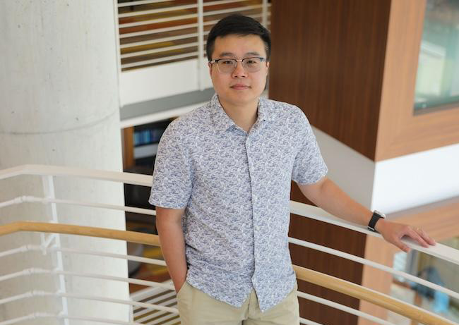 Person wearing glasses and a patterned short‑sleeve shirt, standing and leaning on a railing inside a modern building.