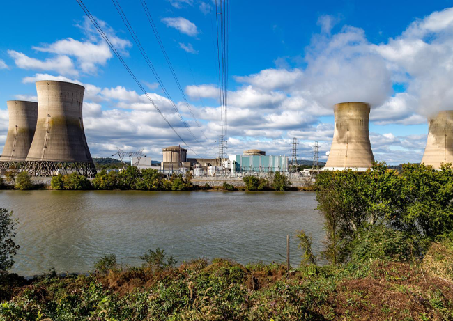 Nuclear power plant with multiple cooling towers beside a river, power lines overhead, and vegetation in the foreground under a partly cloudy sky.