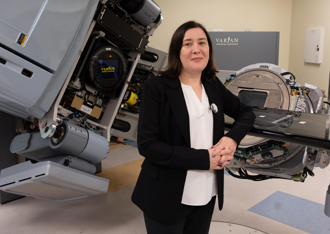 Person standing in a laboratory beside large nuclear research equipment and instrumentation, with mechanical components and control systems visible.