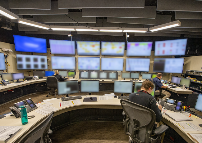 Control room with rows of monitors on a wall and multiple workstations arranged in a semicircle with staff seated at computers.