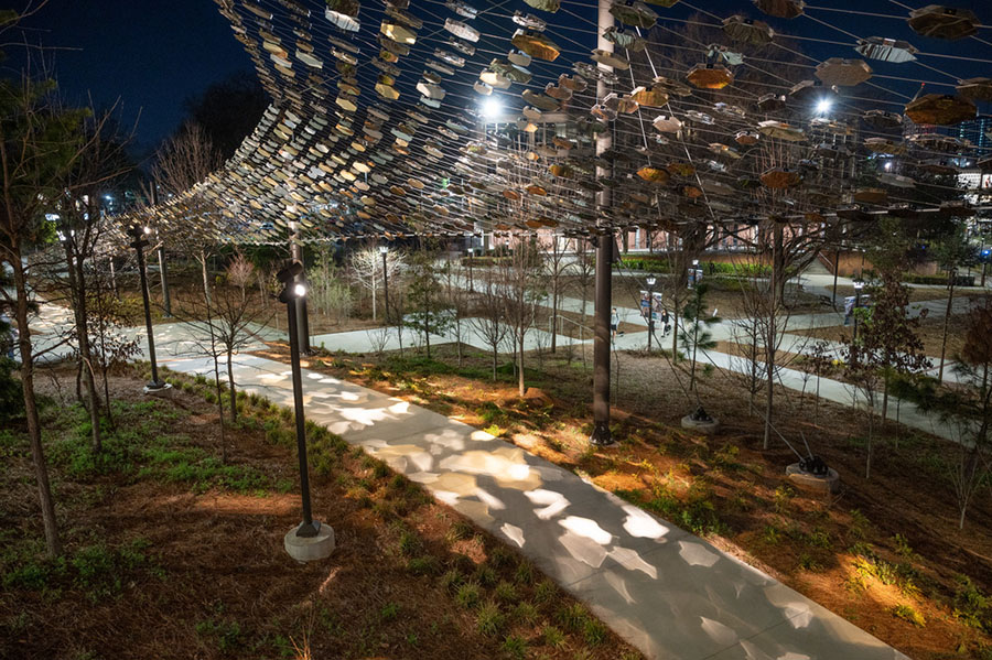 Campus walkway at night under the Pathway of Progress installation, with overhead mirrored panels, trees, and patterned light reflections.