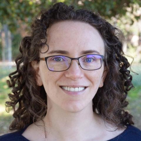 A woman with dark curly hair and glasses wearing a dark blouse smiles for a photo with grass and trees in the background.