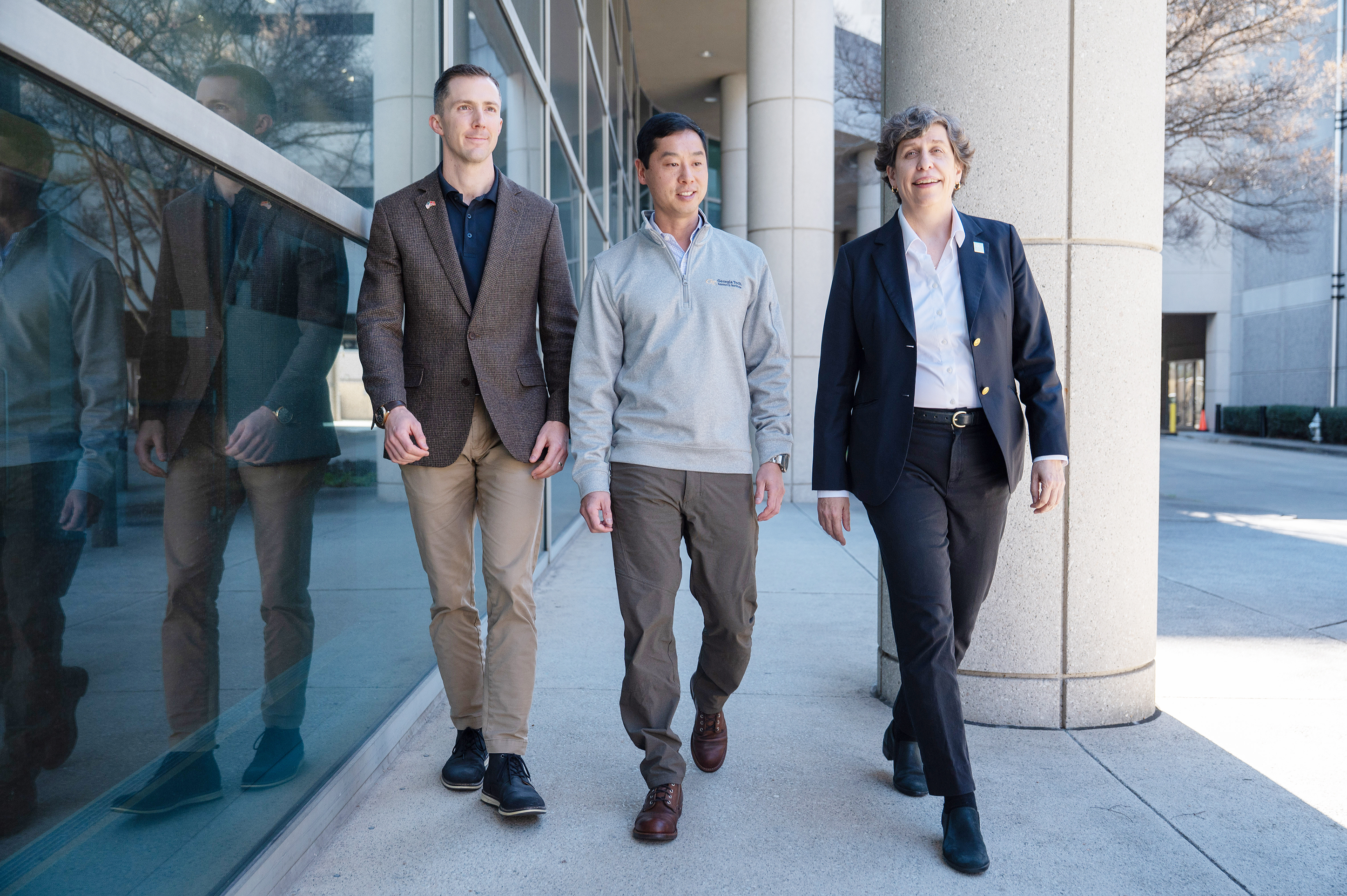 Three people walking together outside a modern building with large glass windows and concrete columns.