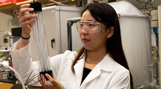 A woman with dark hair holds a glass tube filled with white fibers. 