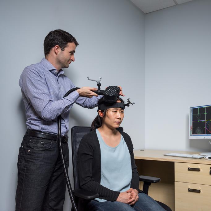 Researcher adjusting a device on another person’s head in a lab, with a computer displaying brain imaging data and a mounted camera in the background.