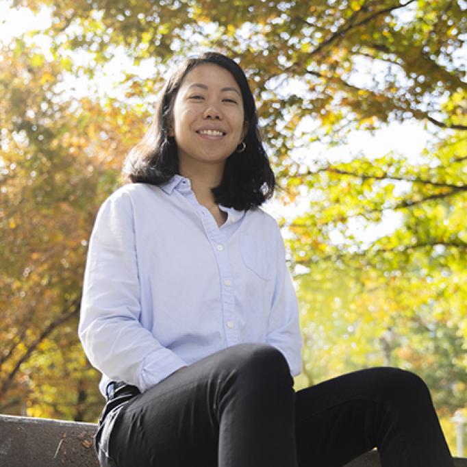 Person sitting outdoors on stone steps wearing a light blue button-up shirt and black pants, with autumn trees and sunlight in the background.
