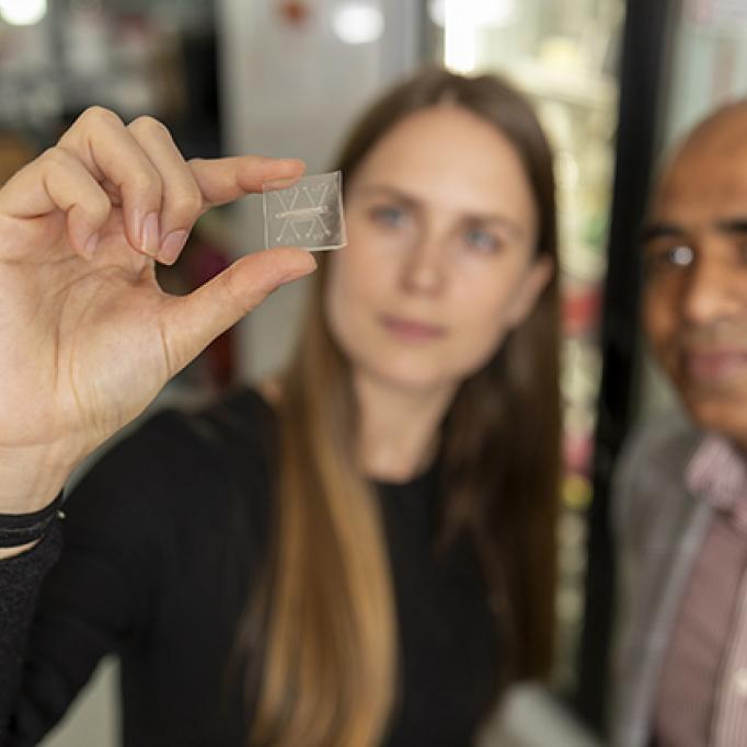 Person holding a small transparent microchip between fingers, with another individual standing nearby in a laboratory setting.