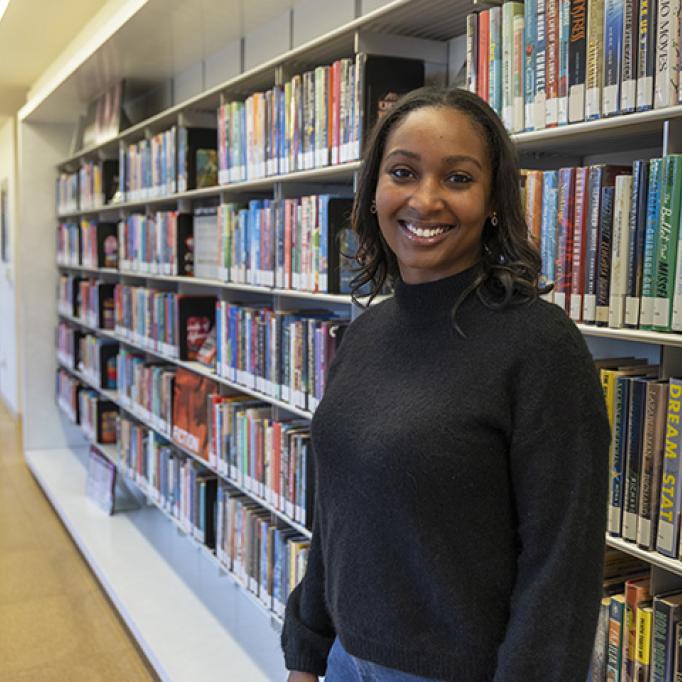 Person standing in a library aisle next to shelves filled with colorful books, wearing a dark sweater and jeans.