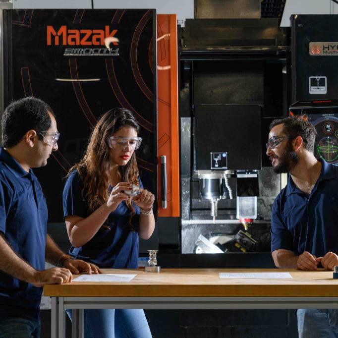 Three individuals standing at a workbench in front of a large Mazak hybrid manufacturing machine, discussing components and technical drawings.