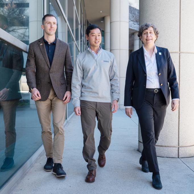 Three people walking together outside a modern building with large glass windows and concrete columns.
