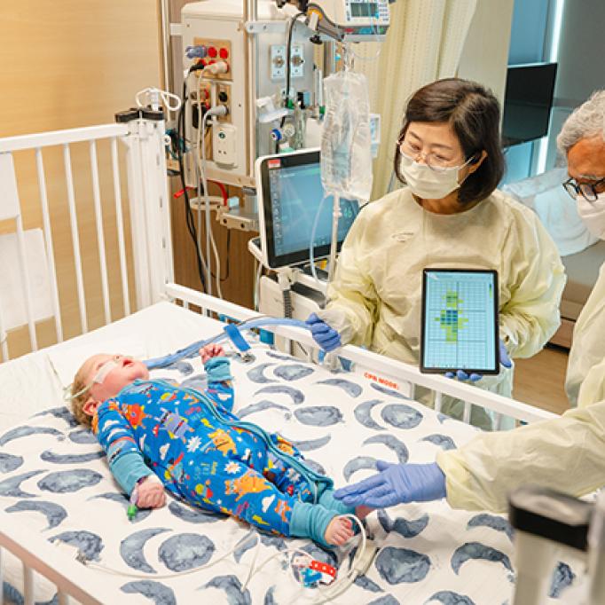 Two adults wearing protective gowns and gloves stand beside a hospital crib, using a tablet device while examining an infant lying on the mattress as medical equipment and monitors surround the crib.