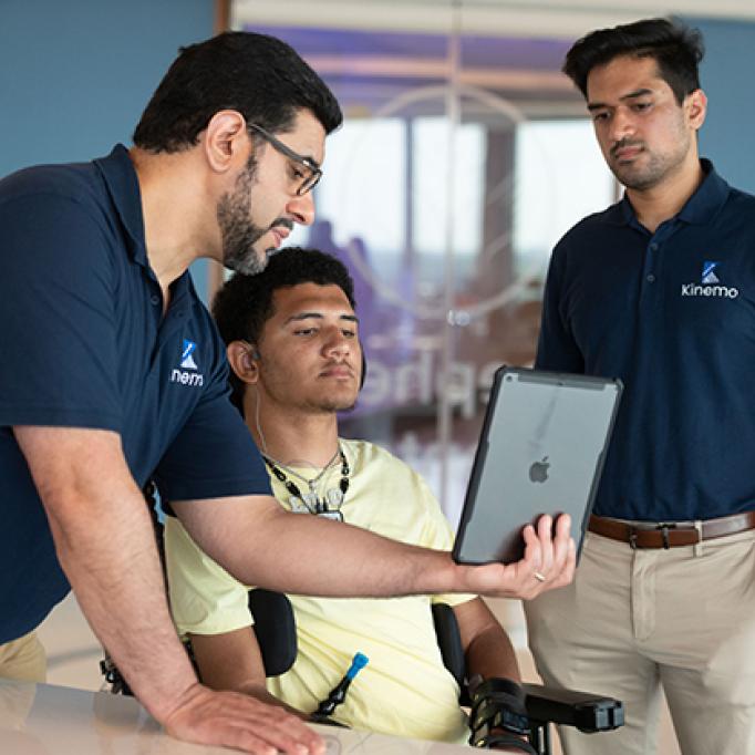Three people review a tablet together in a lab setting, with one seated individual using a powered wheelchair and assistive device.