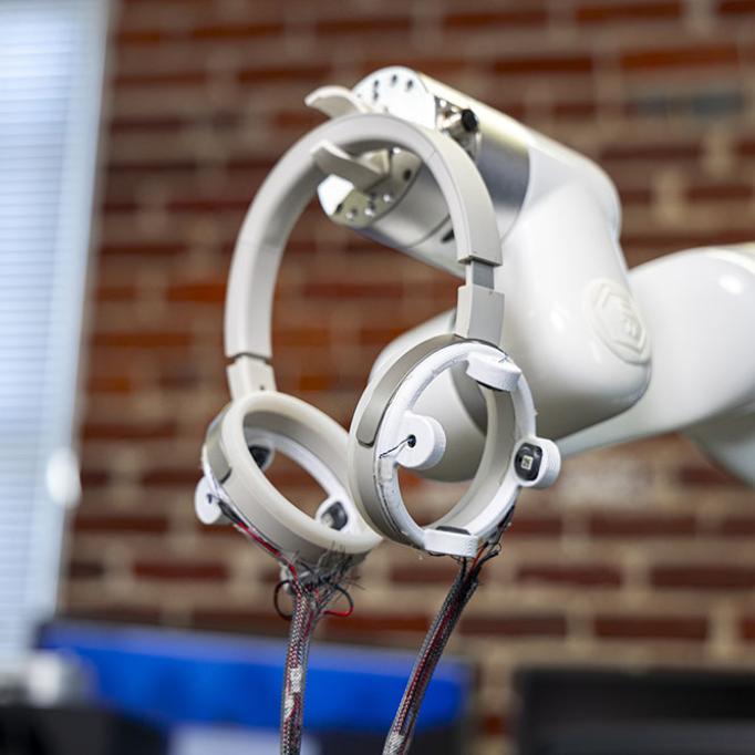 Robotic arm holding circular sensor devices with exposed wiring in a lab setting with a blurred brick wall background.