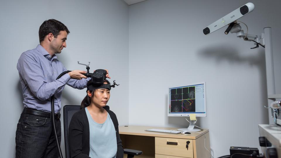 Researcher adjusting a device on another person’s head in a lab, with a computer displaying brain imaging data and a mounted camera in the background.